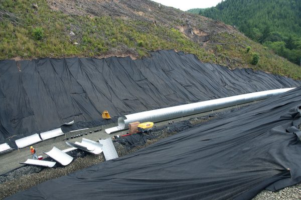 Tubería metálica corrugada de gran diámetro instalada sobre una cama de grava, con el talud de la ladera cubierto de geotextil negro